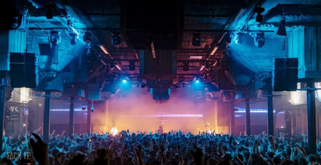 A massive, industrial venue with towering ceilings, thick concrete walls, and a crowd energized by powerful techno beats, all bathed in dynamic lighting.